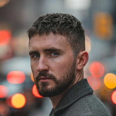 A man with short dark hair and a beard, wearing a dark gray wool coat, standing on a city street at night with blurred car lights in the background.